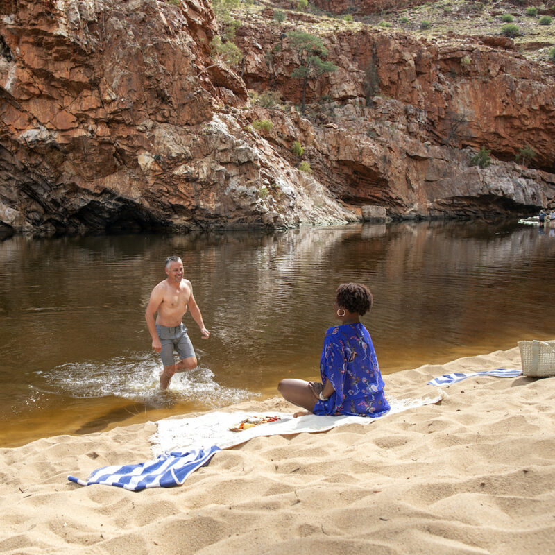 Real Aussie Adventures, Small Group Adventure Tours Australia. West MacDonnell National Park. Woman sitting on the bank while man takes a swim at Ormiston Gorge waterhole Mandatory credit: Tourism NT. Uluru Tours.